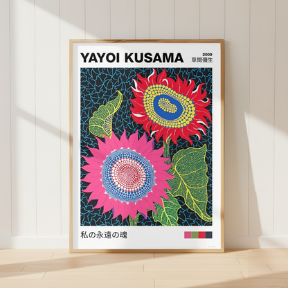 A framed art print of Yayoi Kusama's 1989 sunflower series, leaning against a white paneled wall on a light wood floor. The print features two vibrant sunflowers in pink, red, and green on a dark blue patterned background, with the artist's name 'YAYOI KUSAMA' printed at the top.