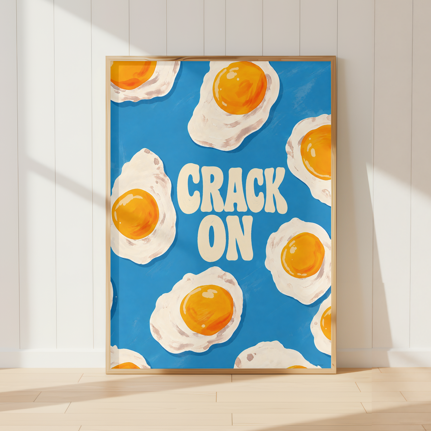 A framed art print featuring a blue background with illustrated sunny-side-up eggs and the words “CRACK ON” in cream lettering, leaning against a white paneled wall on a light wood floor.