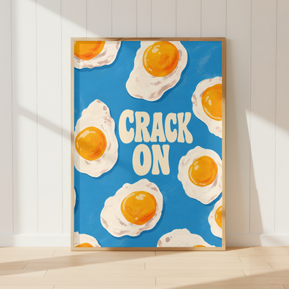 A framed art print featuring a blue background with illustrated sunny-side-up eggs and the words “CRACK ON” in cream lettering, leaning against a white paneled wall on a light wood floor.