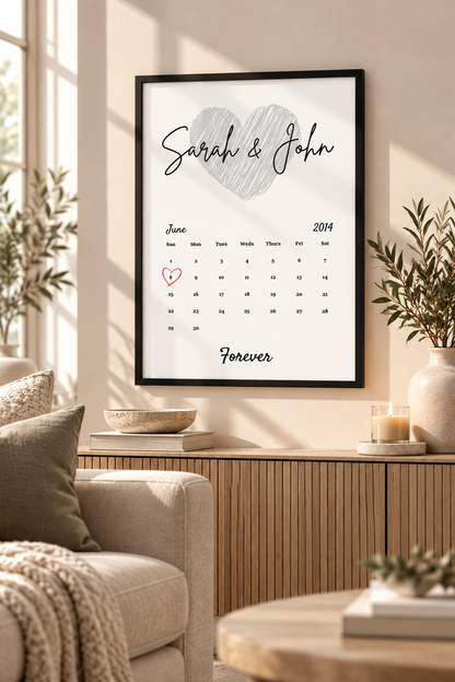 A framed personalized wall calendar print hanging in a styled living room. The print features a large gray heart at the top, the names 'Sarah & John' in script, a June 2014 calendar grid with a small red heart marking the 14th, and the word 'Forever' at the bottom.