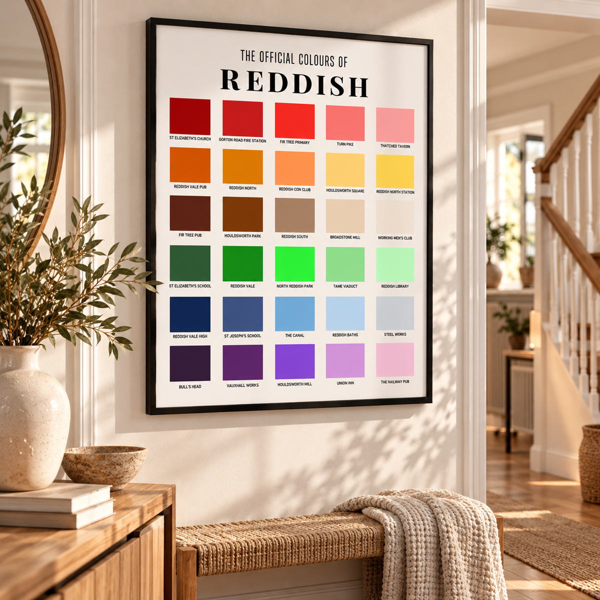 A framed poster titled 'THE OFFICIAL COLOURS OF REDDISH' hangs on a white wall in a bright, modern interior. The poster features a grid of many color blocks ranging from reds and oranges to greens, blues, purples, and pinks, each labeled with a color name.