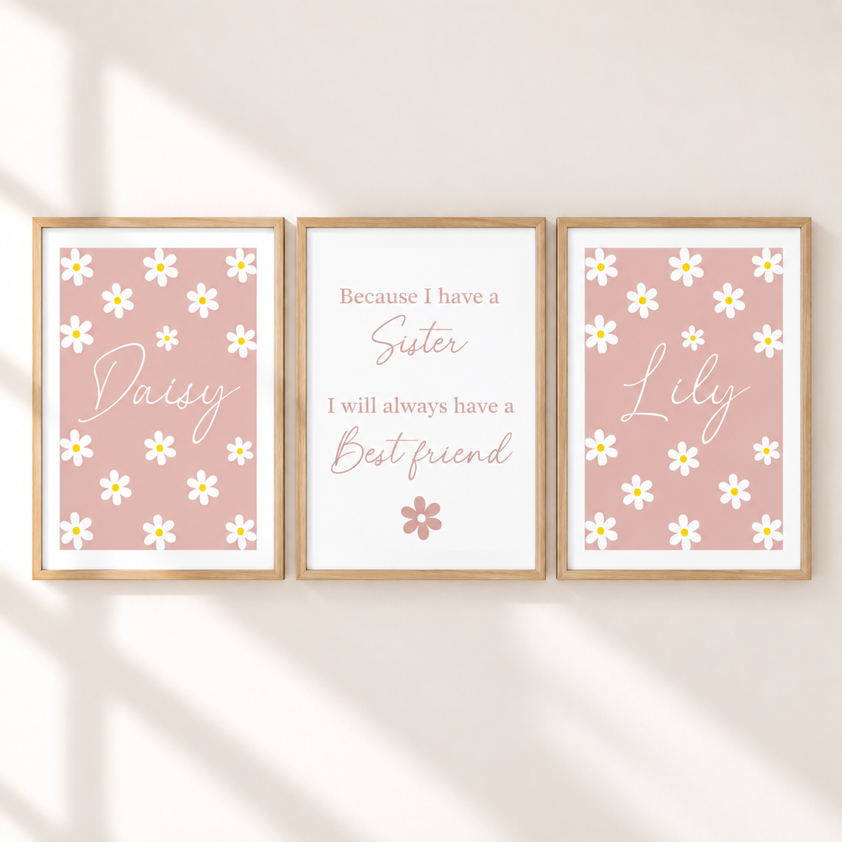 Three framed personalized nursery art prints on a warm beige wall with soft diagonal shadows. The left print says 'Daisy' with white daisies on a dusty pink background, the center print reads 'Because I have a Sister I will always have a Best friend' on a white background, and the right print says 'Lily' with white daisies on a dusty pink background.