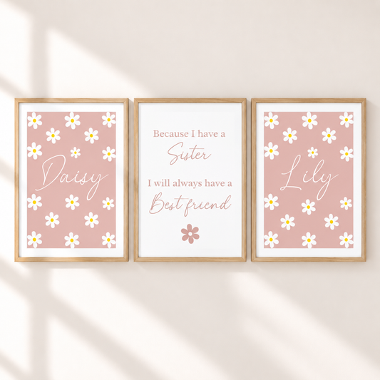 Three framed personalized nursery art prints on a warm beige wall with soft diagonal shadows. The left print says 'Daisy' with white daisies on a dusty pink background, the center print reads 'Because I have a Sister I will always have a Best friend' on a white background, and the right print says 'Lily' with white daisies on a dusty pink background.