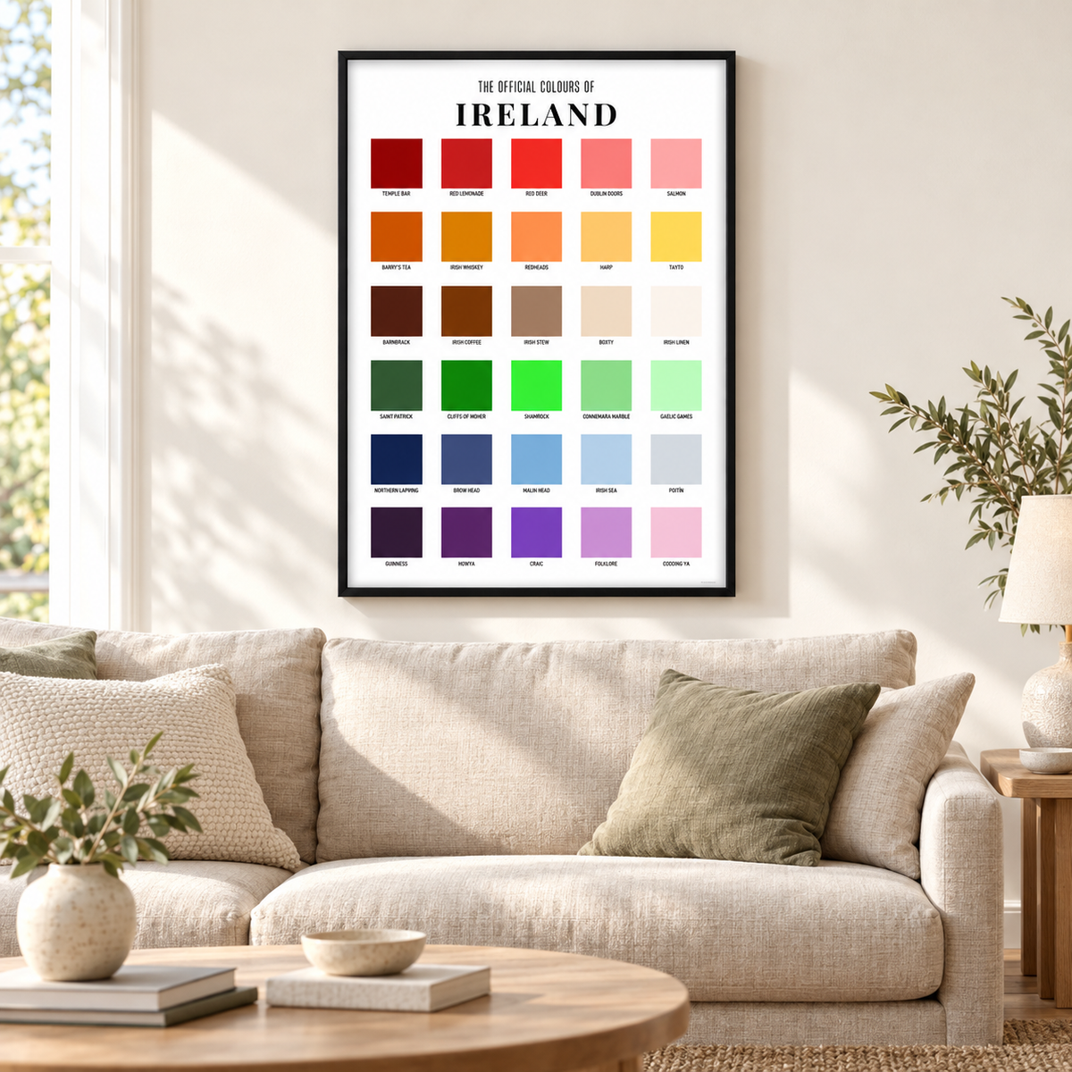 A framed poster titled 'THE OFFICIAL COLOR OF IRELAND' hangs on a beige wall above a light beige sofa in a styled living room. The poster contains a grid of many color swatches representing the Irish flag and landscape, including red, orange, yellow, green, blue, purple, brown, and pink.