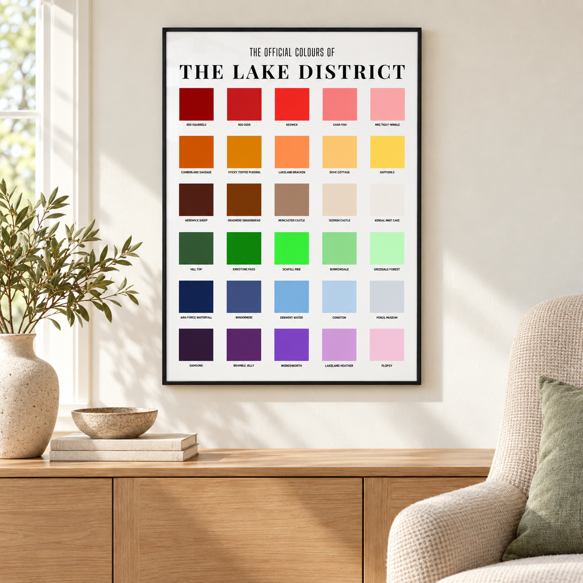 Lake District colour chart print in a thin black portrait frame above an oak sideboard in a bright lounge, featuring 30 colourful squares with local labels.