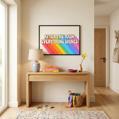 A framed art print with a colorful rainbow background and the text 'AFTER THE RAIN EVERYTHING SHINES' hangs on a beige wall above a light wood console table in a bright, modern room.