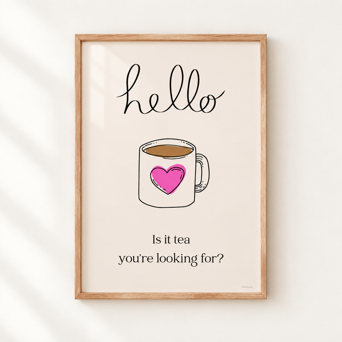 A framed typographic art print featuring a hand-drawn coffee mug with a pink heart, the word 'hello' at the top, and the phrase 'Is it tea you're looking for?' at the bottom, shown hanging on a light wall.