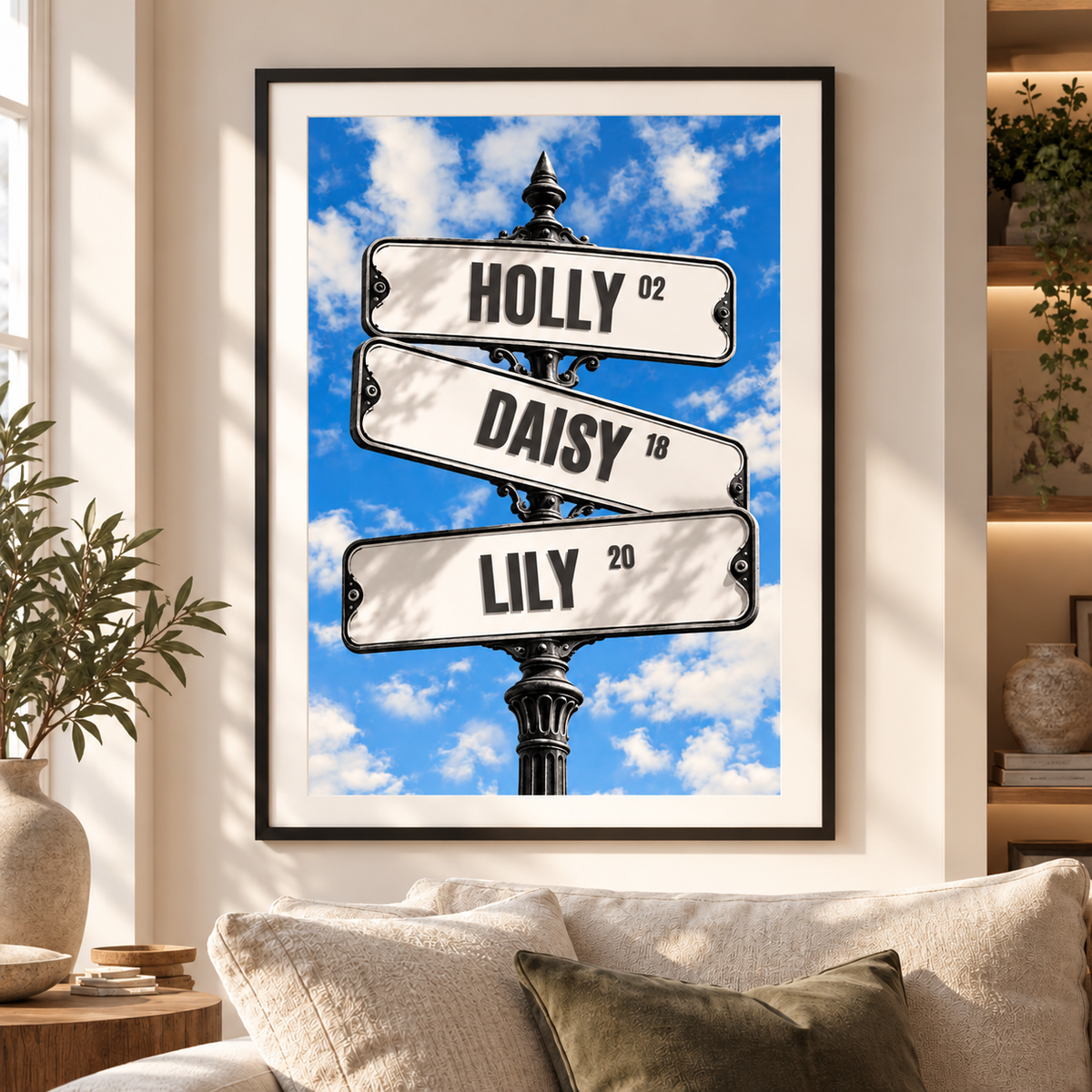 Framed wall art hanging above a sofa in a living room, showing three white street-sign style labels reading HOLLY, DAISY, and LILY against a bright blue sky with clouds.