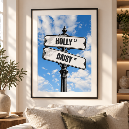 A framed wall art print hanging above a sofa in a living room. The print shows a street sign post with two signs reading HOLLY '92 and DAISY '18 against a blue sky with white clouds.