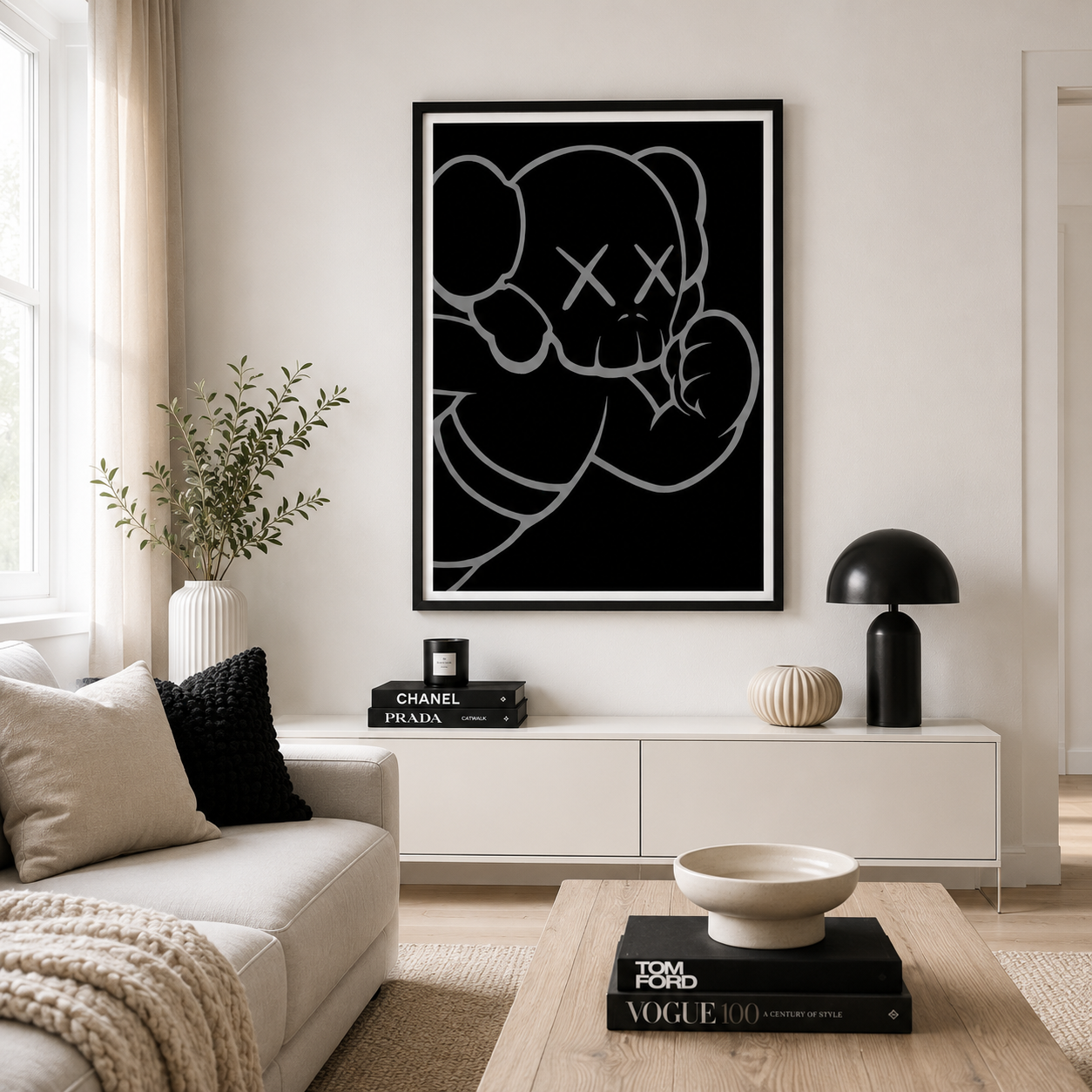 A framed black and white art print of a cartoon bear with an X for a eye hangs on a light beige wall above a white console table in a modern living room setting.