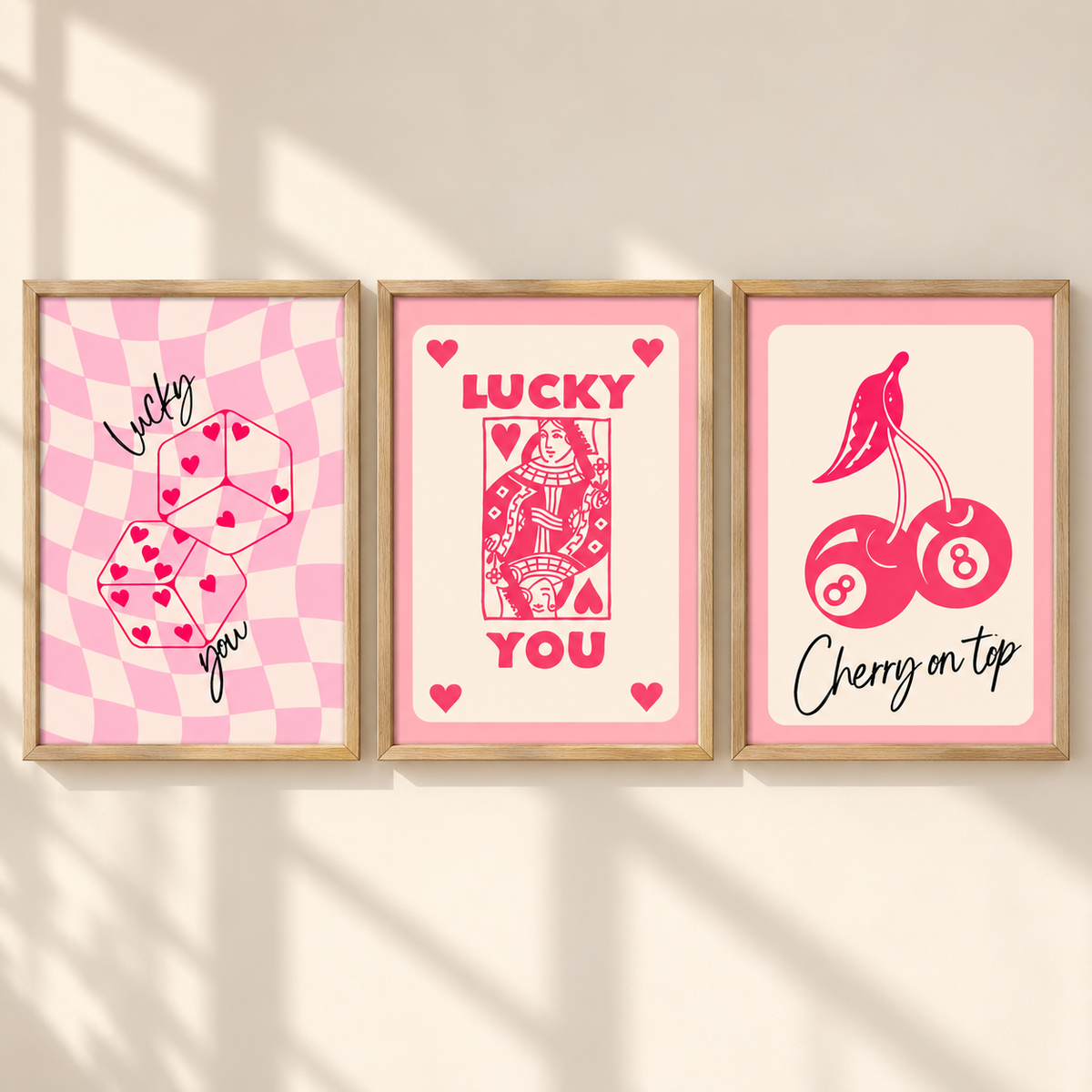 Three framed art prints with pink and red designs hang on a beige wall. The left print shows pink checkered dice with hearts and the words 'Lucky you'; the center print shows a red playing card with 'LUCKY YOU'; the right print shows pink cherries and billiard balls with 'Cherry on tap'.