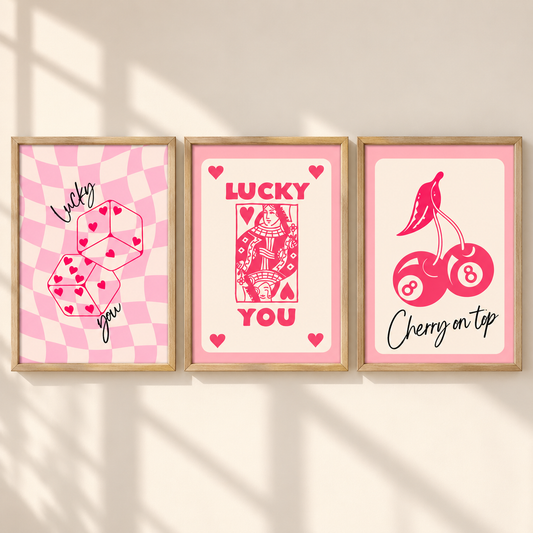 Three framed art prints with pink and red designs hang on a beige wall. The left print shows pink checkered dice with hearts and the words 'Lucky you'; the center print shows a red playing card with 'LUCKY YOU'; the right print shows pink cherries and billiard balls with 'Cherry on tap'.