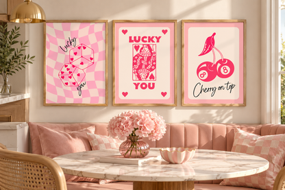 Three framed pink art prints hang above a blush pink sofa in a styled dining or living room. The prints feature pink-and-white checkerboard designs with dice, a playing card reading 'LUCKY YOU,' and cherries with the text 'Cherry on top.