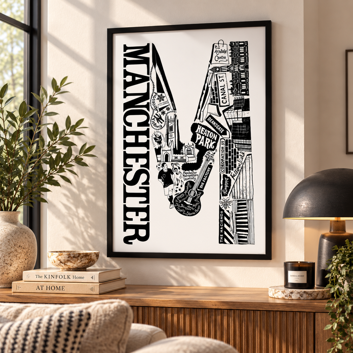 A framed black and white Manchester cityscape art print hangs on a beige wall above a wooden console table in a styled living room setting.