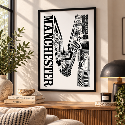 A framed black and white Manchester cityscape art print hangs on a beige wall above a wooden console table in a styled living room setting.