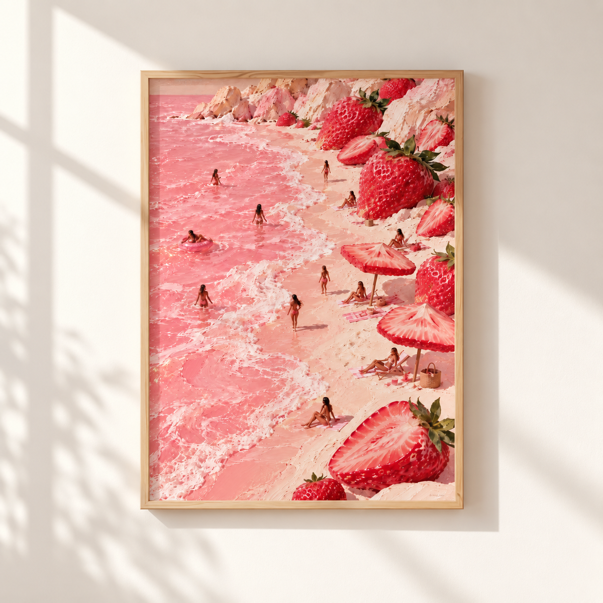 A framed art print depicting a whimsical pink beach scene with strawberries, beach umbrellas, and tiny people in the water, hung on a light beige wall with soft window shadows.