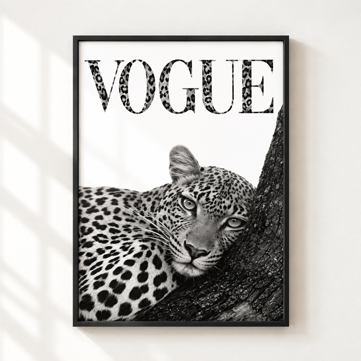 A framed black and white art print featuring a leopard resting on a tree trunk with the word 'VOGUE' in leopard-print lettering, displayed on a light wall with soft window shadow patterns.