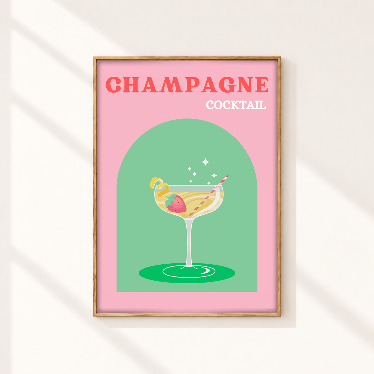 A framed art print featuring a retro-style illustration of a champagne cocktail on a pink background with the text 'CHAMPAGNE COCKTAIL' in coral and white lettering. The print is housed in a light wood frame and displayed on a warm off-white wall with soft diagonal shadows.