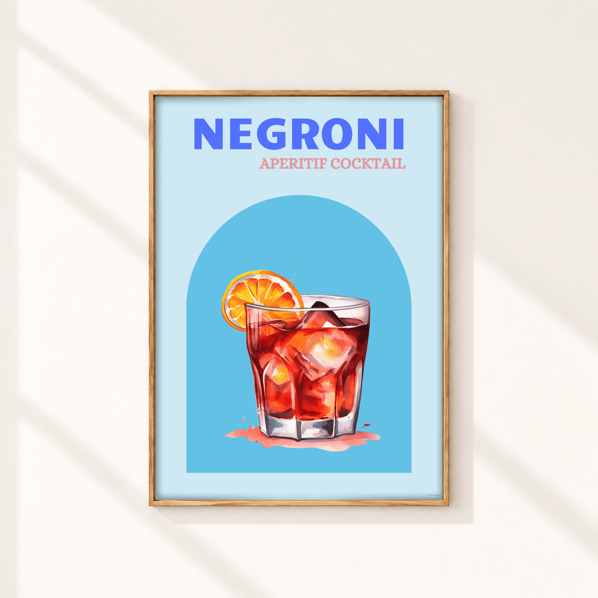 A framed art print featuring a stylized illustration of a Negroni aperitif cocktail with a blue background and a light wood frame, displayed on a warm off-white wall with soft diagonal shadows.