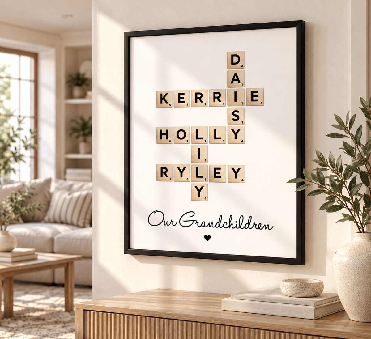 A framed personalized Scrabble-style wall print hangs on a beige wall in a bright living room. The black frame contains wooden letter tiles spelling 'DAD', 'MOTHER', 'PUPIL', 'SISTER', 'RYLEYS', and 'Our Grandchildren' with a small heart below.
