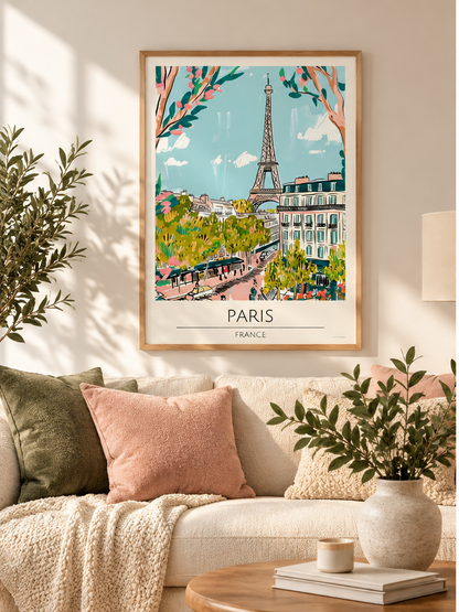 A framed Paris art print hangs on a beige wall above a cream sofa with pink and green pillows in a styled living room. The poster features a colorful illustration of the Eiffel Tower, Paris streets, and trees, with the text 'PARIS FRANCE' at the bottom.