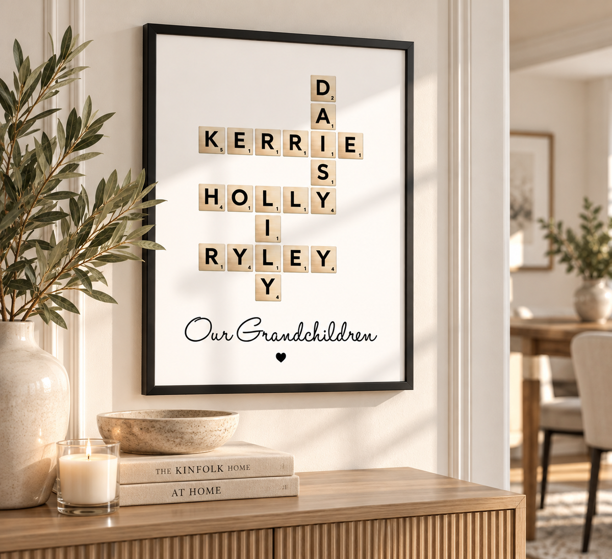 A framed personalized Scrabble-style print hangs on a wall above a wooden console table. The white print features crossword tiles spelling names, with the words 'DAIS', 'KERRIE', 'HOLLY', 'I', 'RYLEY', and 'Our Grandchildren' written below in black script.