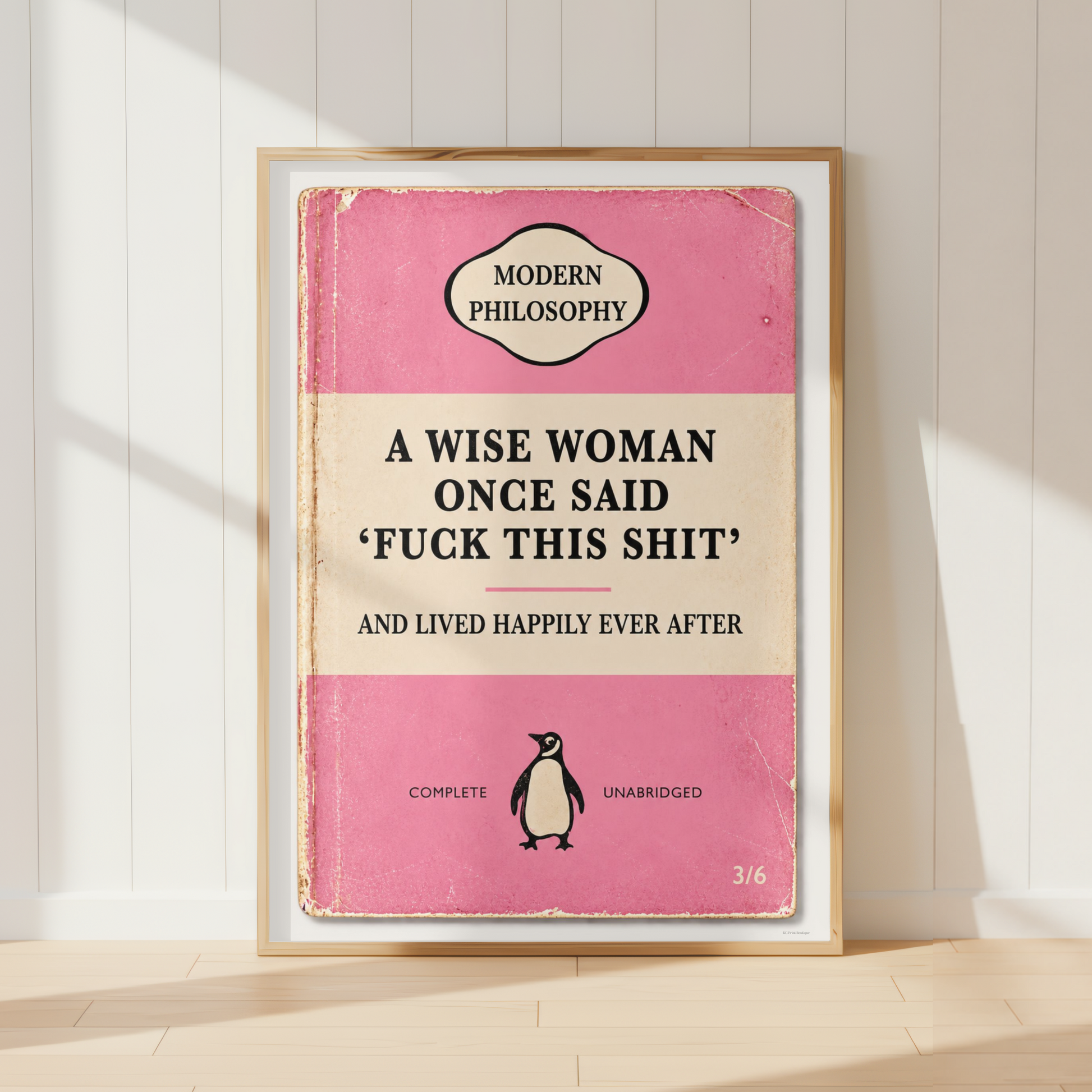 A framed book cover print leaning against a white paneled wall on a light wood floor. The cover is pink and cream with the title 'A WISE WOMAN ONCE SAID 'FUCK THIS SHIT' AND LIVED HAPPILY EVER AFTER' and a small penguin illustration.