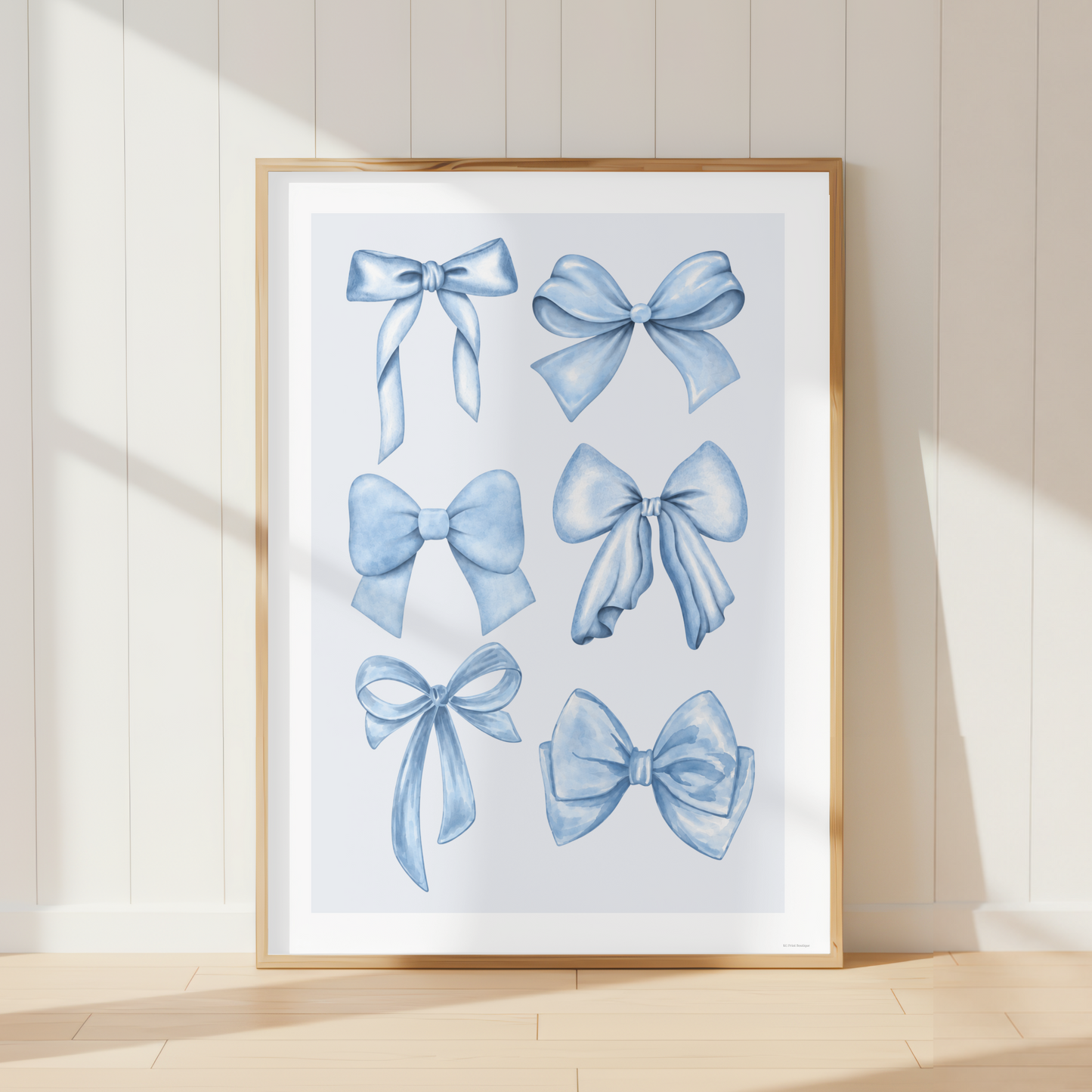 A framed art print featuring six watercolor-style blue ribbon bows, leaning against a cream paneled wall on a light wood floor.