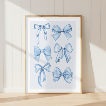 A framed art print featuring six watercolor-style blue ribbon bows, leaning against a cream paneled wall on a light wood floor.
