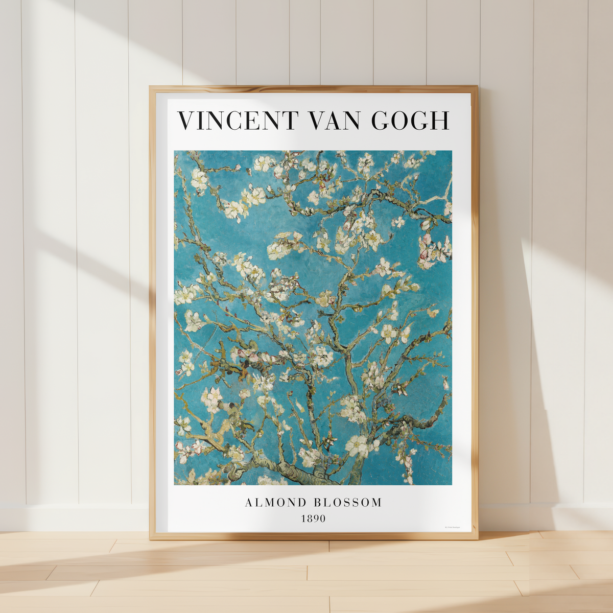 A framed art print of Vincent van Gogh's 'Almond Blossom' leaning against a white paneled wall on a light wood floor, with soft sunlight casting shadows.