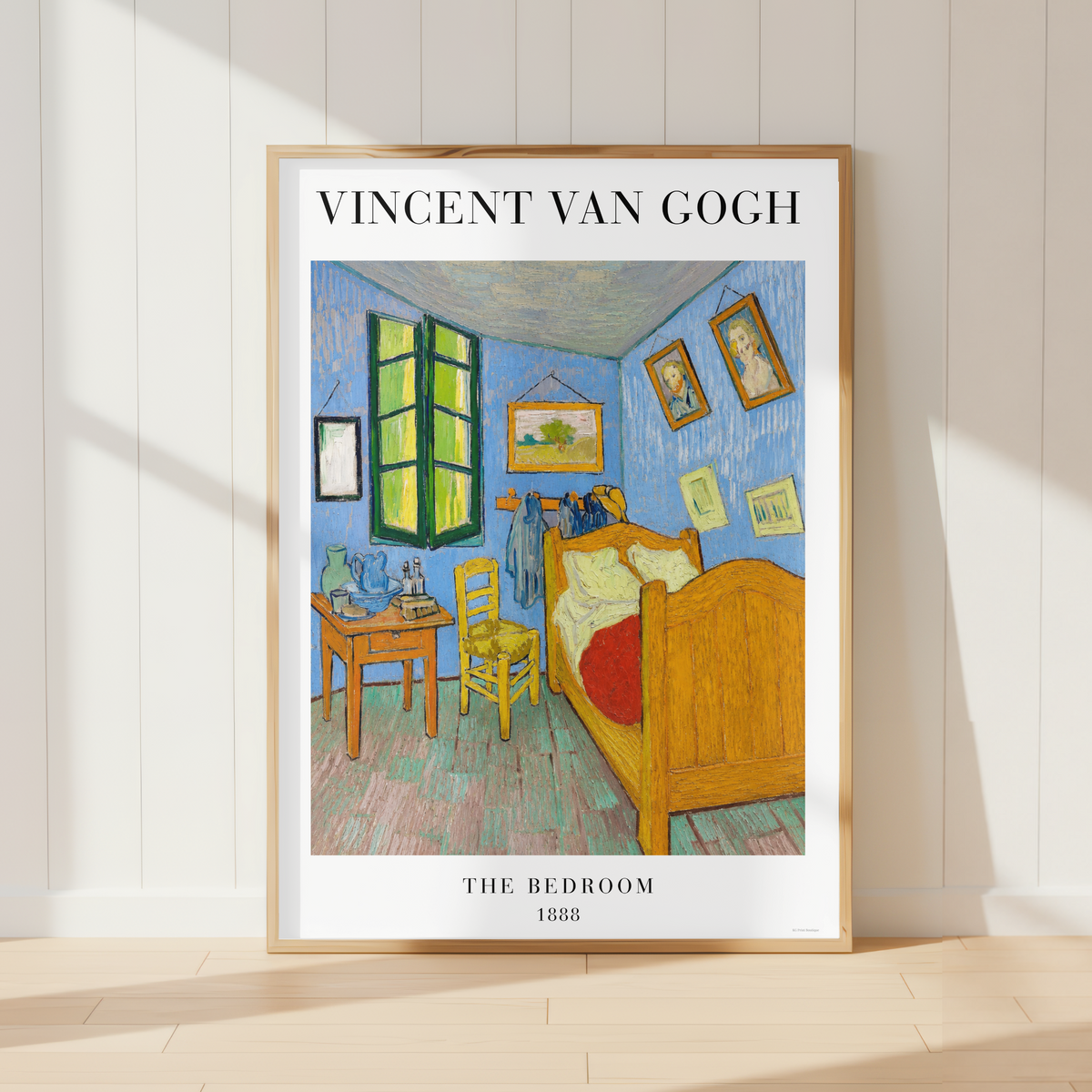 A framed art print of Vincent van Gogh's 'The Bedroom' leaning against a white paneled wall on a light wood floor, with soft sunlight casting shadows.
