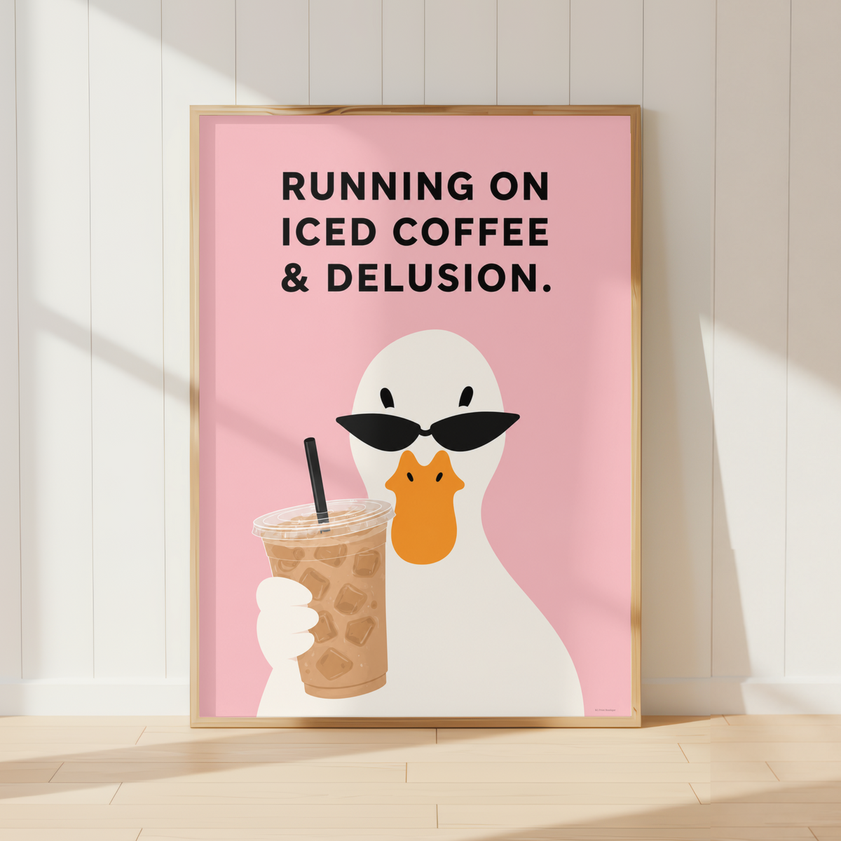 A framed art print leaning against a white paneled wall on a light wood floor. The print features a white duck wearing black sunglasses and holding an iced coffee cup against a pink background, with the text 'RUNNING ON ICED COFFEE & DELUSION.