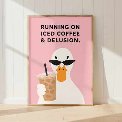 A framed art print leaning against a white paneled wall on a light wood floor. The print features a white duck wearing black sunglasses and holding an iced coffee cup against a pink background, with the text 'RUNNING ON ICED COFFEE & DELUSION.