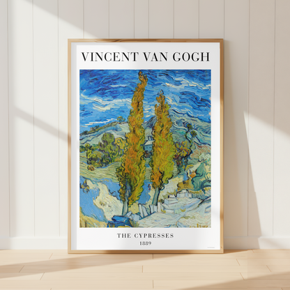 A framed art print of Vincent van Gogh's 'The Cypress Trees at Arles 1909' leaning against a white paneled wall on a light wood floor, with soft sunlight casting shadows.