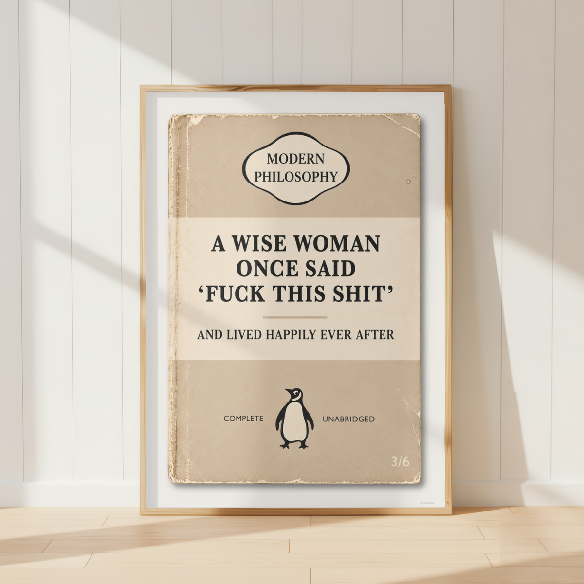 A framed book-style poster leaning against a white paneled wall on a light wood floor. The poster has a vintage beige cover design with the text 'A WISE WOMAN ONCE SAID 'FUCK THIS SHIT' AND LIVED HAPPILY EVER AFTER' and a small Penguin logo near the bottom.