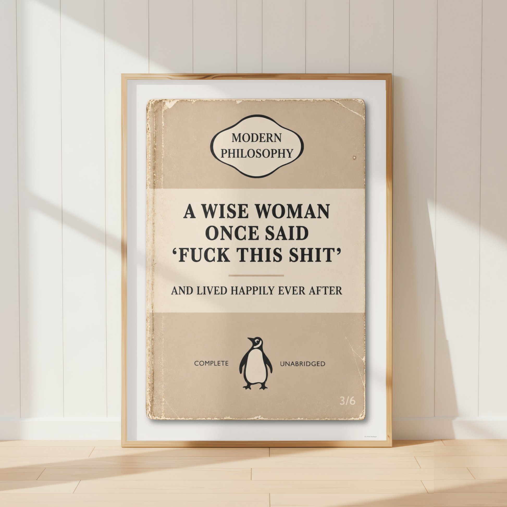 A framed book-style poster leaning against a white paneled wall on a light wood floor. The poster has a vintage beige cover design with the text 'A WISE WOMAN ONCE SAID 'FUCK THIS SHIT' AND LIVED HAPPILY EVER AFTER' and a small Penguin logo near the bottom.