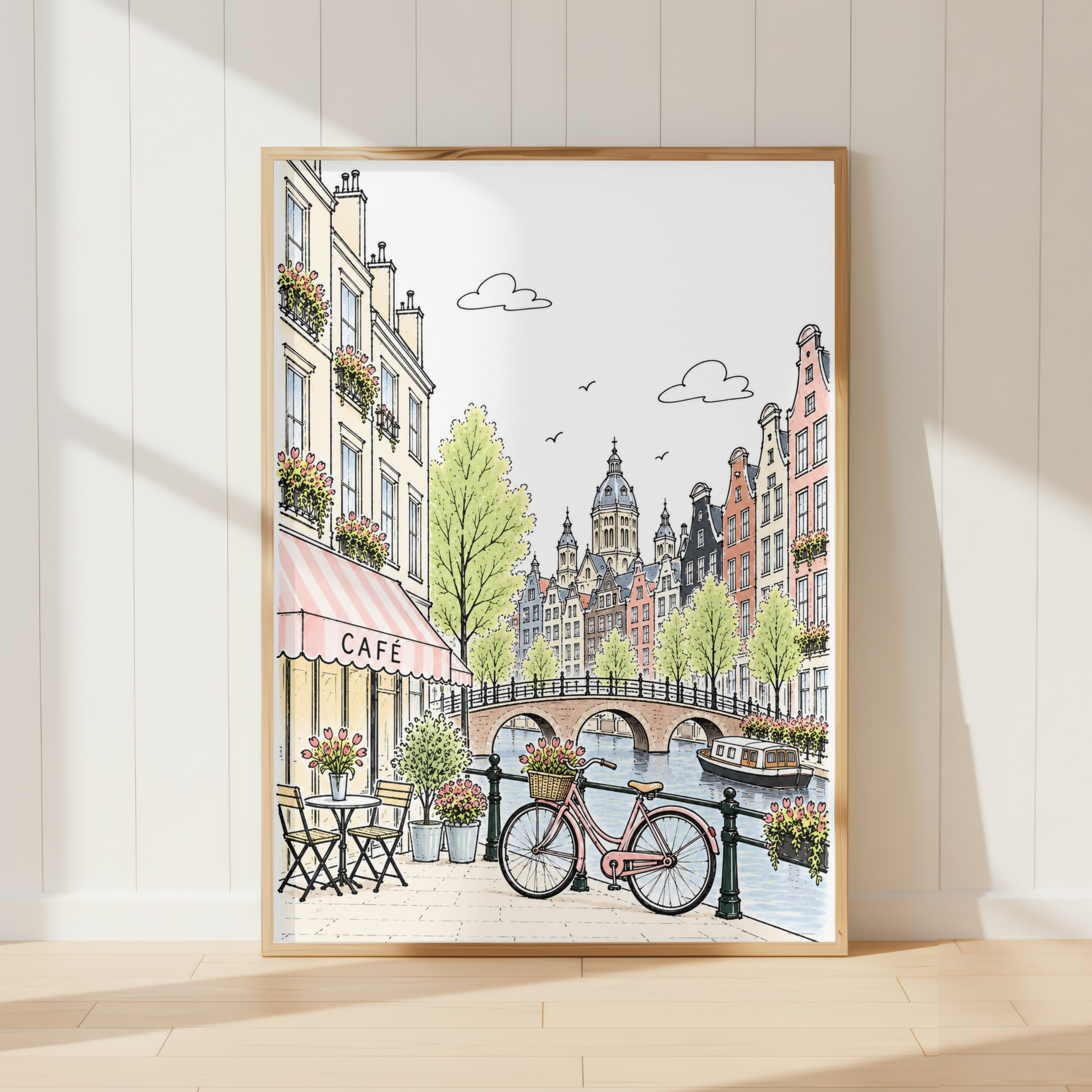 A framed art print depicting a charming canal-side city street with a bicycle, cafe seating, and historic buildings, leaning against a white paneled wall on a light wood floor.