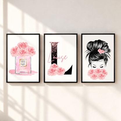 Three framed wall art prints hang on a light beige wall with soft window shadows. The left print shows pink roses in a perfume bottle labeled Chanel; the center print features a black glittery letter L with pink script reading Louise and pink flowers at the bottom; the right print shows a black-and-white illustrated woman with a pink flower in her hair and pink flowers in front of her face.