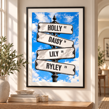 A framed vertical street-sign style wall art print hangs on a beige wall above a wooden console. The artwork shows four white signboards reading HOLLY, DAISY, LILY, and RYLEY against a bright blue sky with white clouds, in a black frame.