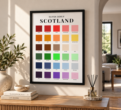 A framed poster titled 'The Official Colours of Scotland' hangs on a beige wall in a styled living room. The poster features a grid of colorful color swatches with labels, surrounded by home decor including a plant, books, and a reed diffuser.