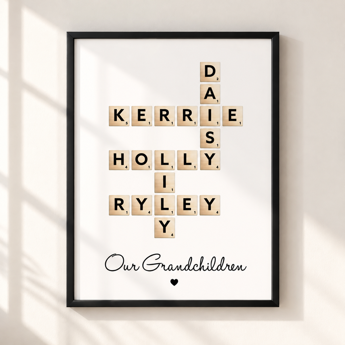 A framed personalized Scrabble-style print hanging on a light wall, with wooden letter tiles spelling names across a crossword layout and the phrase 'Our Grandchildren' in black script below. The frame is black and the wall behind it is warm off-white with soft window shadows.