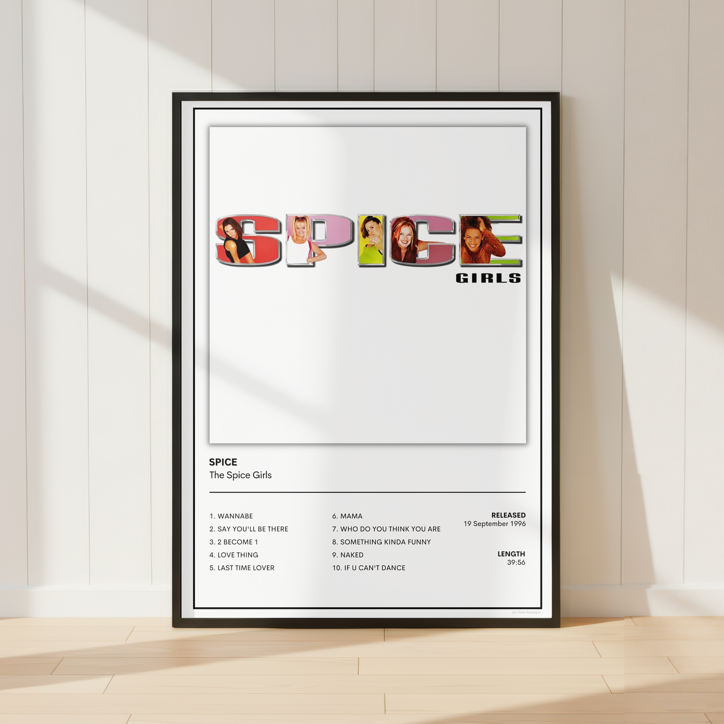 A framed Spice Girls 'Spice' music poster leaning against a white paneled wall on a light wood floor, with soft sunlight casting shadows across the scene.