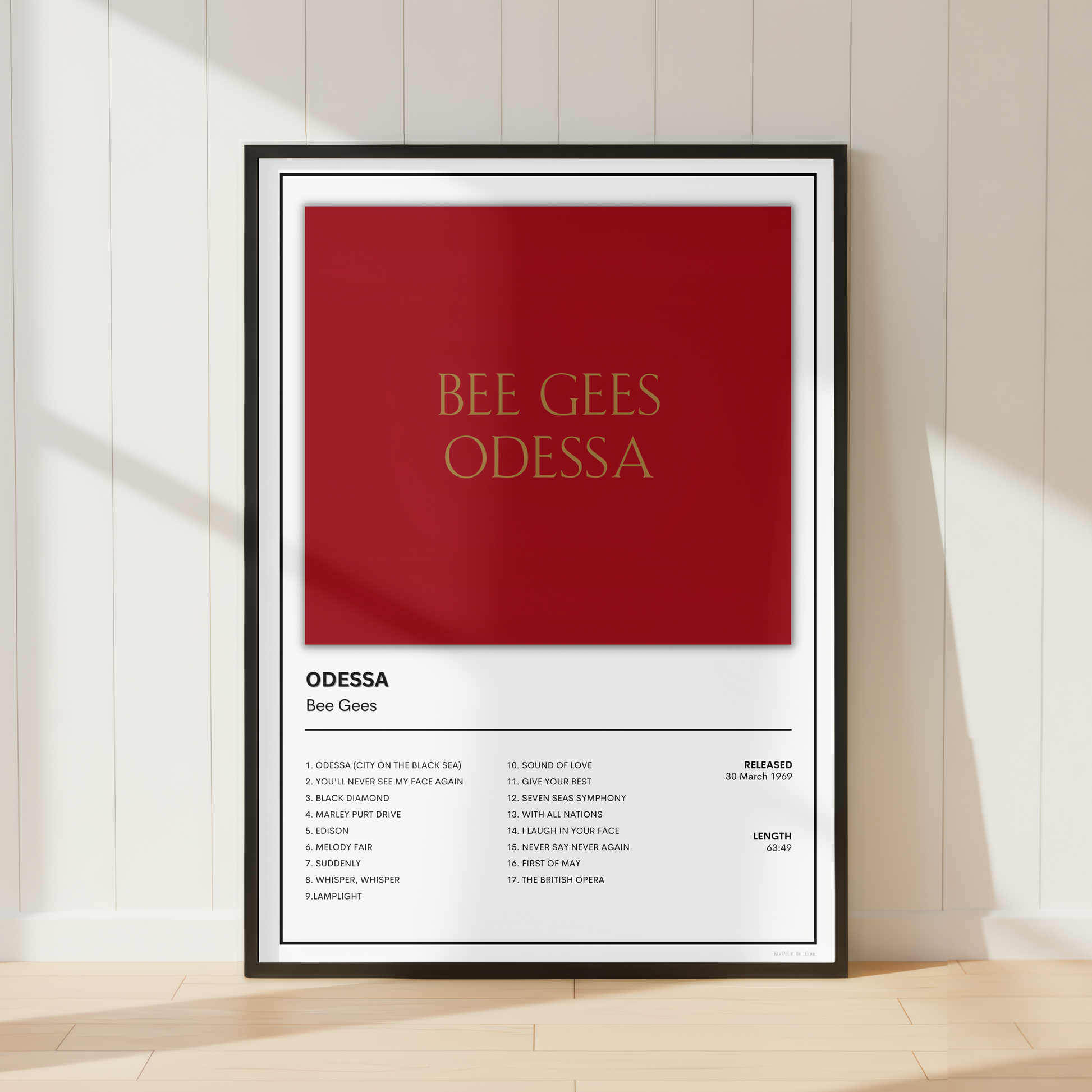 A framed poster print featuring a red and white design with the text 'BEE GEES ODESSA' in gold, displayed against a white paneled wall on a light wood floor.