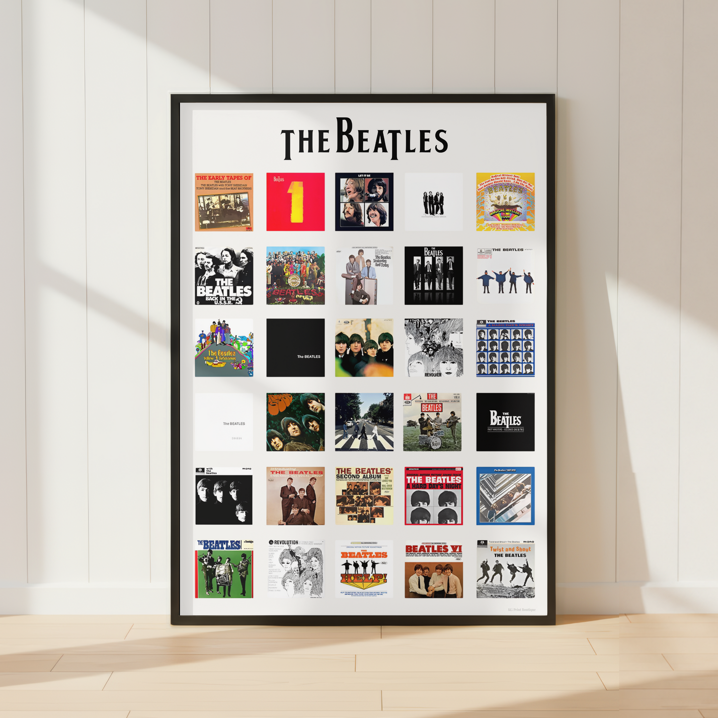 A framed poster featuring a grid of The Beatles album covers under the large 'THE BEATLES' title, displayed against a white paneled wall on a light wood floor with soft sunlight and shadows.