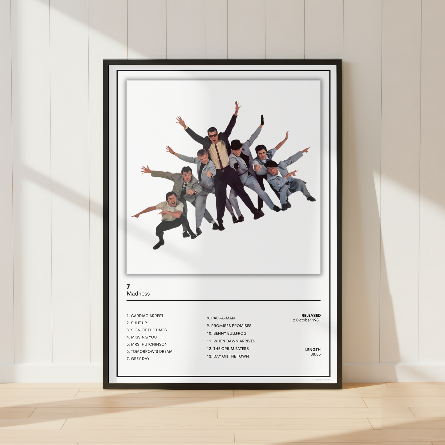 A framed music poster of the band Madness '7' leaning against a white paneled wall on a light wood floor, with soft window shadows cast across the scene.