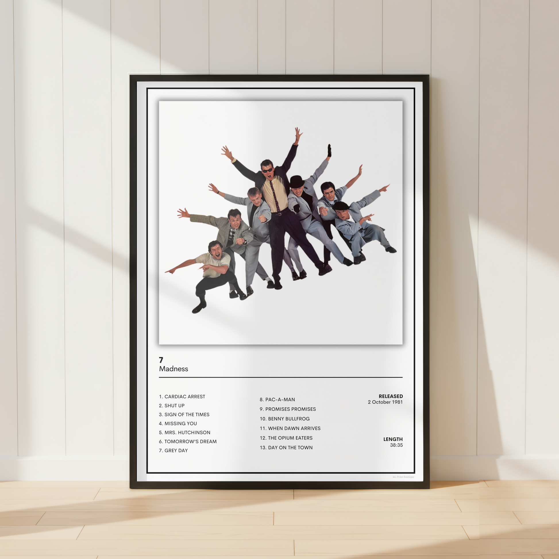 A framed music poster of the band Madness '7' leaning against a white paneled wall on a light wood floor, with soft window shadows cast across the scene.