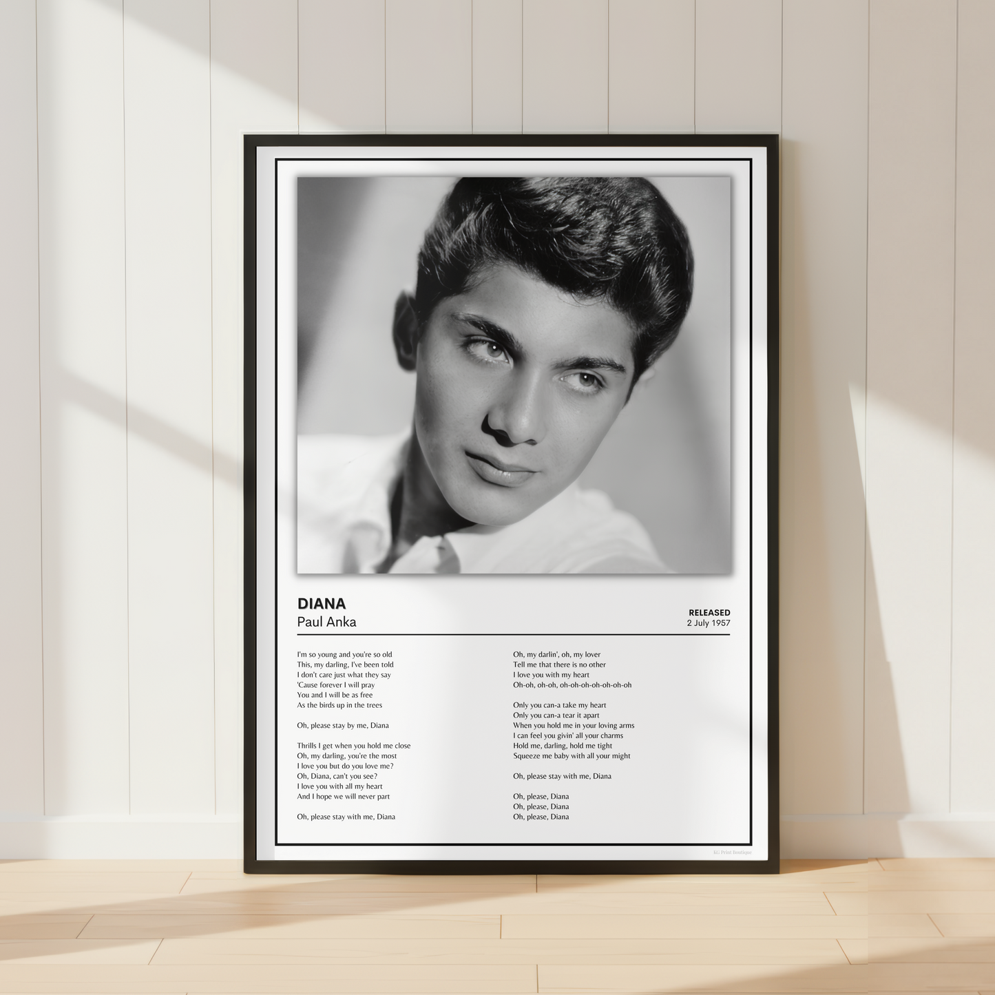 A framed black-and-white poster print leaning against a light wood-paneled wall on a pale wood floor. The poster features a portrait of Paul Anka with the text 'DIANA Paul Anka' and song lyrics printed below.