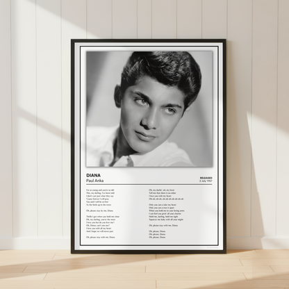 A framed black-and-white poster print leaning against a light wood-paneled wall on a pale wood floor. The poster features a portrait of Paul Anka with the text 'DIANA Paul Anka' and song lyrics printed below.