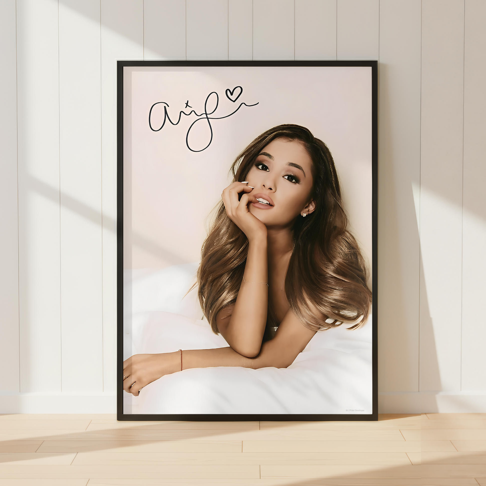 A framed portrait print of Ariana Grande leaning against a white paneled wall on a light wood floor, with soft sunlight casting shadows across the scene.