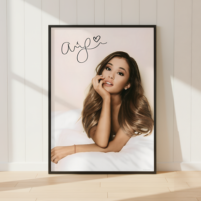 A framed portrait print of Ariana Grande leaning against a white paneled wall on a light wood floor, with soft sunlight casting shadows across the scene.